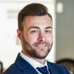 Businessman leader in modern office with businesspeople working Caucasian businessman leader looking at camera in modern office with multi-ethnic businesspeople working at the background. Teamwork concept. Young man with beard wearing blue suit.